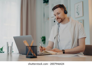 Smiling young man sitting at table and using headset while looking at laptop screen and making notes in notebook, male freelance in headphones having online meeting with colleagues. Freelance concept