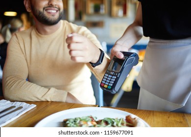 Smiling Young Man With Smartwatch Keeping His Wrist Close To Payment Machine While Using Transfer System To Pay For Meal