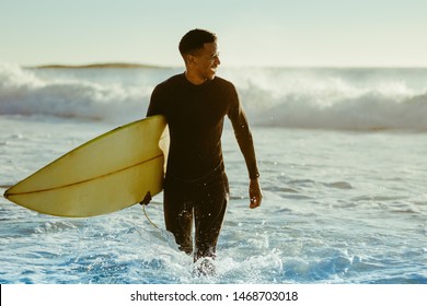 Smiling young man with surfboard on the beach. African male coming out of the ocean after water surfing.