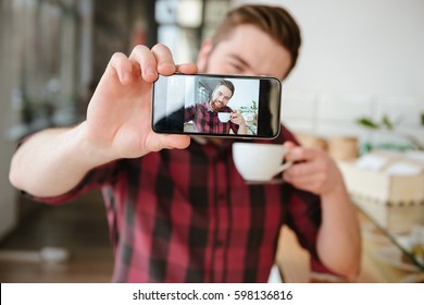 Smiling young man taking selfie and holding cup of coffee while sitting at cafe - Powered by Shutterstock