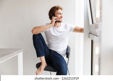 Smiling Young Man Talking On Mobile Phone, Looking At The Window At Home