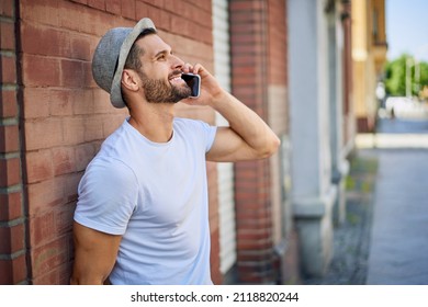 Smiling young man talking on the phone while leaning against a wall in the city - Shutterstock ID 2118820244