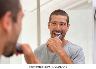 Smiling young man with toothbrush cleaning teeth and looking mirror in the bathroom. Handsome man brushing his teeth in morning in bathroom. Happy guy in pajamas brushing teeth before going to sleep.