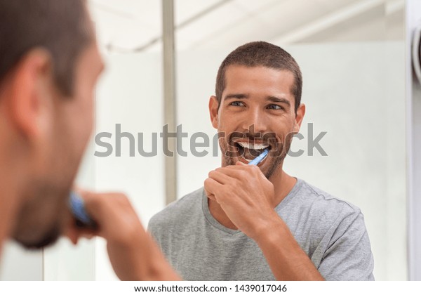 Smiling young man with toothbrush cleaning teeth\
and looking mirror in the bathroom. Handsome man brushing his teeth\
in morning in bathroom. Happy guy in pajamas brushing teeth before\
going to sleep.