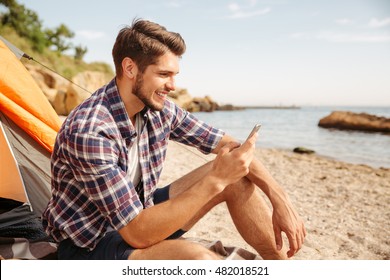 Smiling young man tourist using smartphone sitting in touristic tent at the beach - Powered by Shutterstock