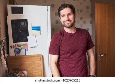 Smiling Young Man In A T-shirt Standing Alone By The Fridge In His Kitchen In The Morning