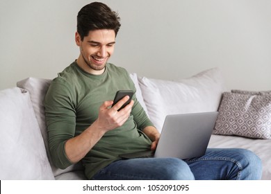 Smiling Young Man Typing On Mobile Phone While Sitting On A Sofa At Home With Laptop Computer