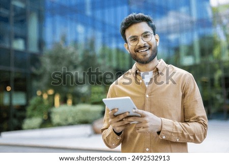 Smiling young man using digital tablet in modern urban environment. Confident individual in casual outfit holding device outdoors. Concept of technology, business, and lifestyle in city setting.