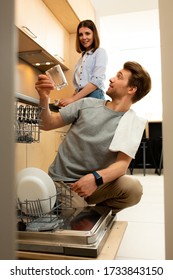 Smiling Young Man Using Dishwasher While Pretty Woman Standing Nearby In The Kitchen At Home