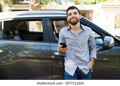 Smiling Young Man Using His Smartphone And Texting While Leaning On A Black Car Outside