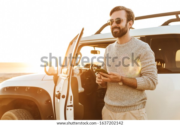 Smiling young man using mobile phone while standing\
at the car at the beach