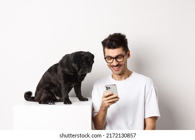 Smiling Young Man Using Smartphone And Sitting Near Dog. Pug Owner Checking Photos On Mobile Phone, White Background