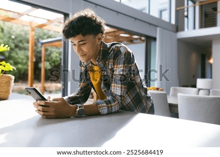 Smiling young man using smartphone while leaning on kitchen counter at home. Technology, lifestyle, communication, millennial, casual, relaxed