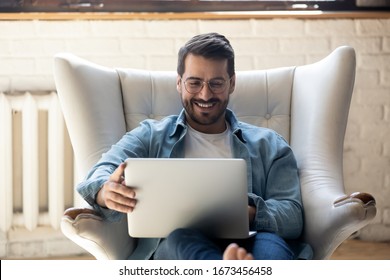 Smiling young man wearing glasses using laptop, sitting in cozy armchair, happy male looking at computer screen, chatting in social network or shopping online, playing game, working at home