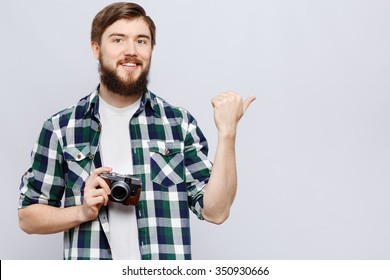 Smiling Young Man, Wearing On White T-shirt And Blue Checked Shirt, Is Showing Something By His Hand And Holding Camera, On White Background, In Studio, Waist Up