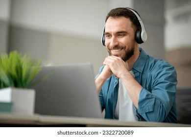 Smiling Young Man In Wireless Headphones Watching Webinar On Laptop, Happy Millennial Male Enjoying Online Education And Distance Learning, Sitting At Desk And Looking At Computer Screen, Free Space - Powered by Shutterstock