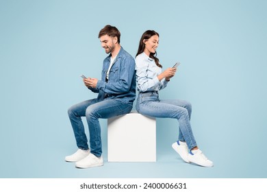 Smiling young man and woman dressed in casual denim outfits are focused on their smartphones while sitting back-to-back against a blue backdrop - Powered by Shutterstock