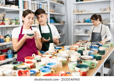 Smiling young man and women in pinafores examining handmade jars of potter-earth material in art studio - Powered by Shutterstock