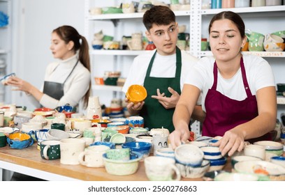 Smiling young man and women in pinafores examining handmade jars of potter-earth material in art studio - Powered by Shutterstock