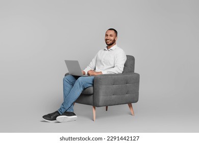 Smiling young man working with laptop in armchair on grey background