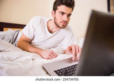 Smiling Young Man Is Working With Laptop, Lying On Bed.