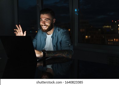 Smiling Young Man Working On Laptop Computer While Sitting At The Table Indoors At Night, Waving Hand