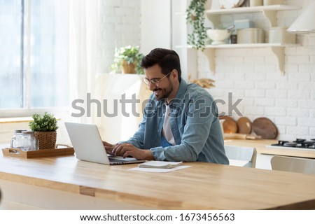Smiling young man working on laptop in modern kitchen, checking email in morning, writing message in social network, happy young male using internet banking service, searching information