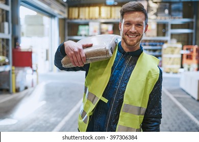 Smiling young man working in a warehouse standing with a bag of product over his shoulder grinning happily at the camera, close up view