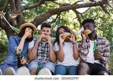 Smiling Young Men And Women Drinking Beer And Eating Burgers During Picnic At Summer Garden. Four Diverse Friends Spending Free Time Together On Fresh Air.