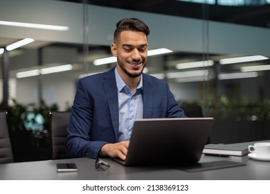 Smiling young middle eastern businessman sitting at worktable at modern office, typing on computer keyboard, sending emails to his business partners, working on marketing research, copy space - Shutterstock ID 2138369123