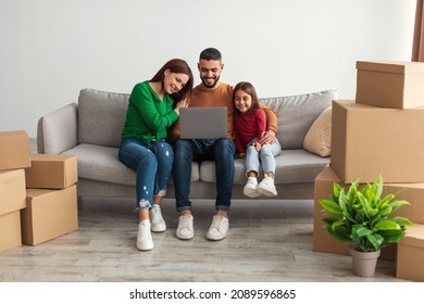 Smiling Young Middle Eastern Family Using Pc Laptop Choosing New Room Design, Sitting On Sofa With Carton Boxes And Plants. People Planning Interior Change Of Apartment, Moving And Buying Furniture