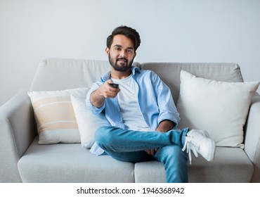 Smiling Young Middle Eastern Guy Watching TV At Home. Millennial Arab Man Relaxing On Couch In Living Room And Switching Channels With Remote Controller, Enjoying Domestic Weekend, Copy Space