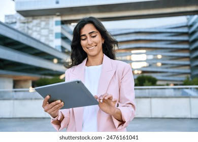 Smiling young middle eastern Israel businesswoman using tablet pc application for online remote work standing at office business building outdoors. Indian or arabic 30s woman holding digital computer - Powered by Shutterstock