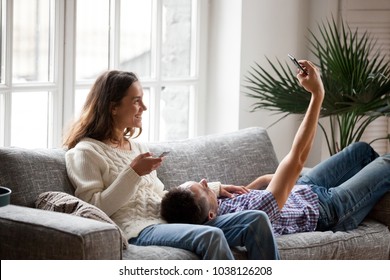 Smiling Young Millennial Couple Having Fun With Smartphones Using Apps At Home Making Photo Or Shooting Blog On Cellphone, Happy Man Taking Selfie On Mobile Phone Relaxing With Girlfriend On Couch