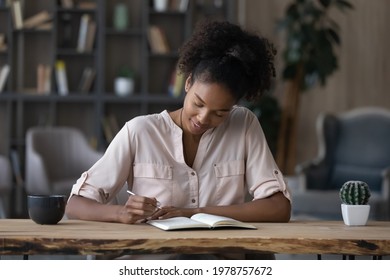 Smiling young mixed race african business woman sitting at table, writing notes in paper planner, managing working issues or making to-do list, planning workday or assignments at modern home office. - Powered by Shutterstock