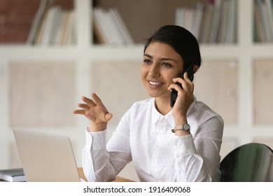 Smiling Young Mixed Race Ethnicity Woman Have Pleasant Cellphone Call Or Conversation. Happy Millennial Indian Female Employee Work On Laptop Talk Speak On Smartphone Consult Client Online.