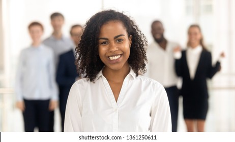 Smiling young mixed race female office worker student intern standing looking at camera with team people at background, happy african american employee business coach millennial professional portrait