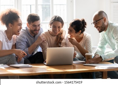 Smiling young mixed race woman showing funny videos on computer to happy multiracial friends, sitting together on cozy couch at home. Overjoyed diverse millennial people having fun in dormitory. - Shutterstock ID 1564175419