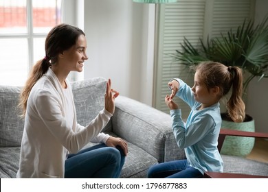 Smiling young mixed race woman teaching little cute girl sign language, showing symbols with fingers. Smiling hearing loss deaf disabled child communicating nonverbal with mommy in living room.
