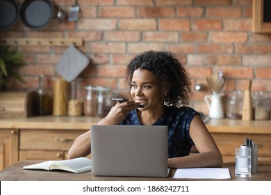 Smiling young mixed race woman dictating audio message in cellphone application, distracted from computer work at home. Happy multiracial lady holding loudspeaker conversation or sending voicemail.