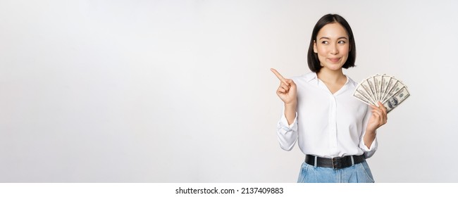 Smiling young modern asian woman, pointing at banner advertisement, holding cash money dollars, standing over white background - Shutterstock ID 2137409883