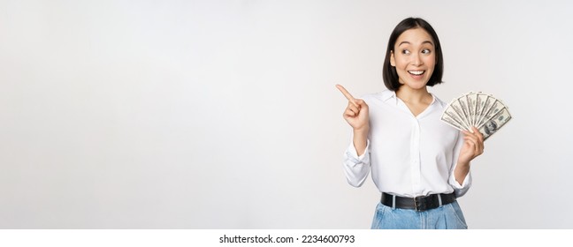 Smiling young modern asian woman, pointing at banner advertisement, holding cash money dollars, standing over white background. - Shutterstock ID 2234600793