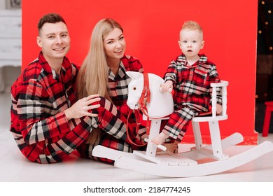 Smiling Young Mom, Dad Are Playing With Baby Boy On Wooden Rocking Horse On The Red Background. Happy Family With Little Child In Red Checked Pyjamas Having Fun Together. Childcare, Childhood