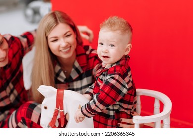 Smiling Young Mom, Dad Are Playing With Baby Boy On Wooden Rocking Horse On The Red Background. Happy Family With Little Child In Red Checked Pyjamas Having Fun Together. Childcare, Childhood