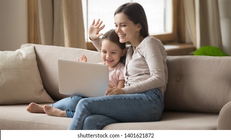 Smiling Young Mom And Funny Little Daughter Sit On Couch Hold Laptop Wave Videochat Talk On Webcam, Happy Preschooler Girl Child And Mother Speak Using Computer Have Online Conversation With Dad