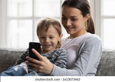 Smiling Young Mom Or Nanny Sit Rest On Sofa Hug Small Boy Child Watch Funny Video On Cellphone Together, Happy Mother Enjoy Time With Little Son Using Smartphone Playing Game In Living Room