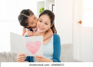 Smiling Young Mom Reading The Mother's Day Gift Card Sitting On The Living Room Sofa With Little Daughter Kiss From Behind At Home.