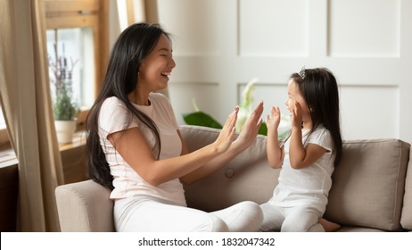 Smiling Young Mother And Adorable Little Daughter Wearing Princess Diadem Clapping Hands, Sitting On Couch, Loving Mum And Cute Toddler Child Girl Having Fun Together, Enjoying Leisure Time