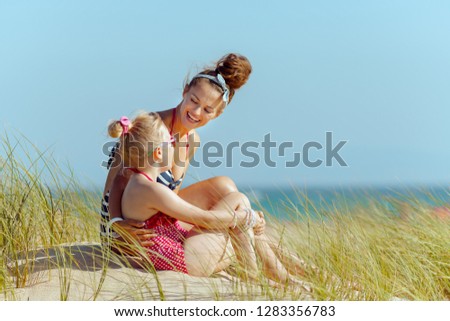 smiling young mother and child in beachwear on the seashore looking at each other. getting vitamin D after long winter months. blond hair daughter in red dotted swimsuit with flowers.