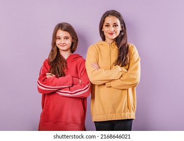 Smiling Young Mother And Cute Daughter Standing Front View With Folded Arms In Fashion Trendy Hoodie Together On Purple Studio Background With Empty Copy Space. Happy Loving Family Portrait